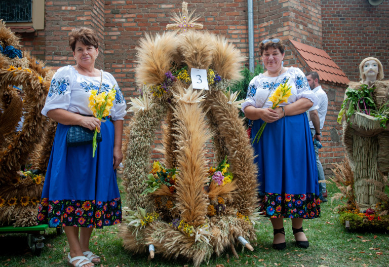 Wieniec ze zbóż i kwiatów stoi przed kościołem. Przy nim stoją dwie kobiety z żółtymi kwiatami.