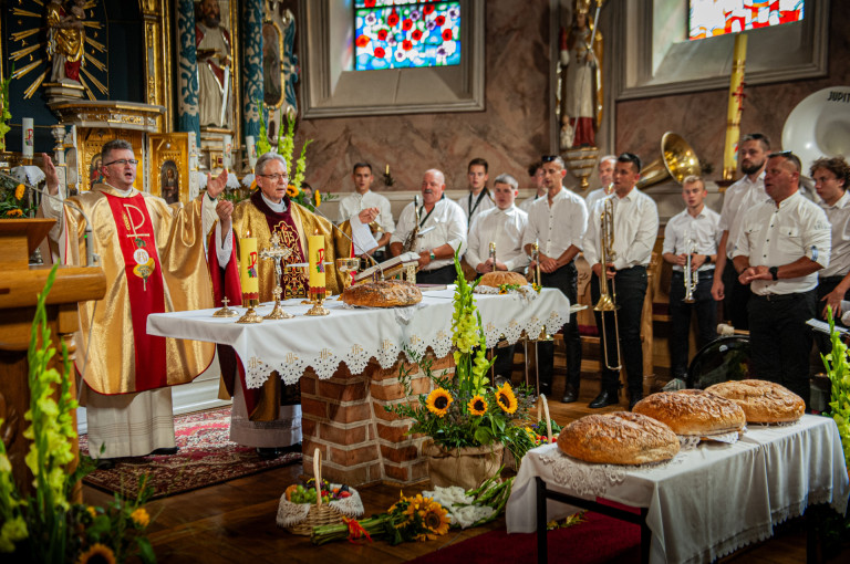 Księża stoją w kościele przy ołtarzu. Obok są członkowie orkiestry z instrumentami.