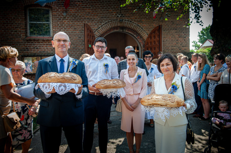 Goście są przed kościołem. Dwóch mężczyzn i dwie kobiety, trzymają trzy bochny chleba.