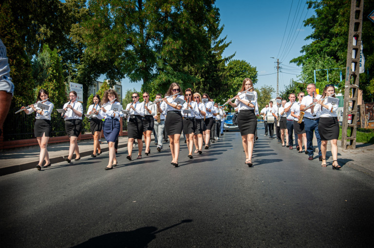 Członkowie orkiestry dętej w biało czarnych strojach idą asfaltową drogą i grają na instrumentach.