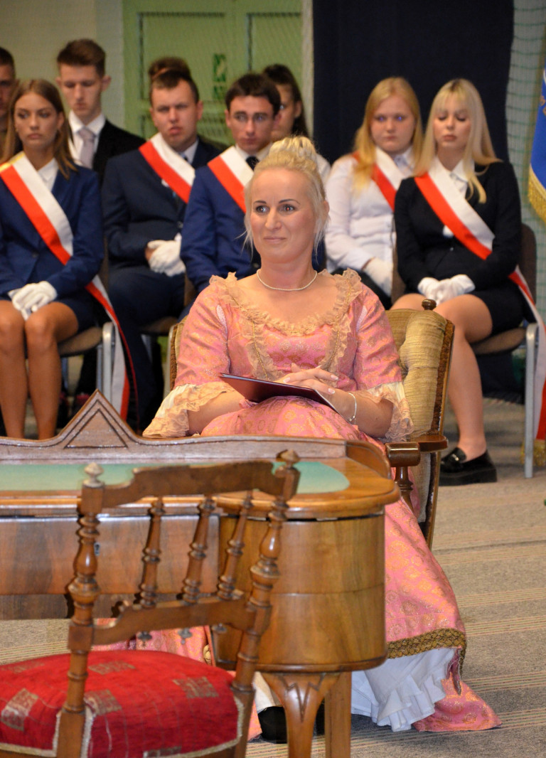 Kobieta w różowej sukni z epoki oświecenia siedzi na fotelu. W tle siedzą uczniowie.
