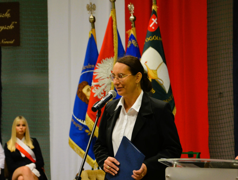 Kobieta w białej koszuli i czarnej marynarce przemawia do mikrofonu na tle sztandarów.
