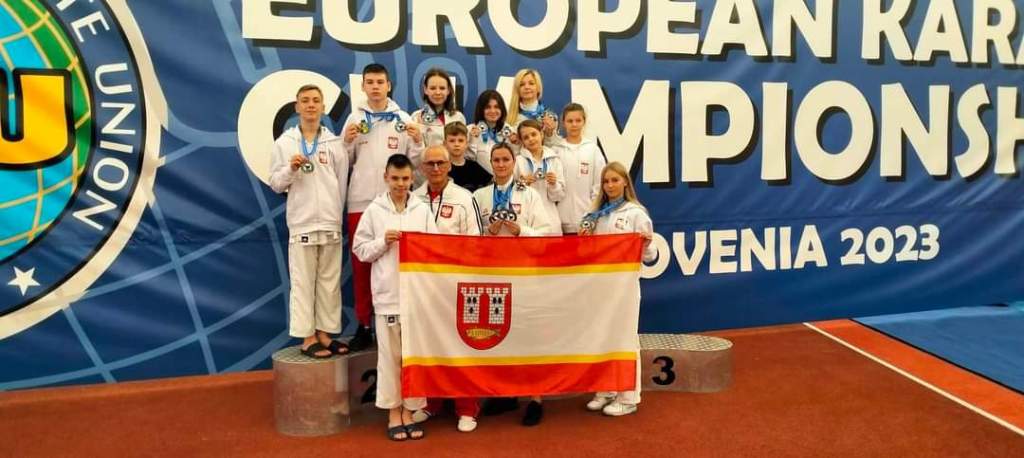 W hali sportowej stoją trener i zawodnicy z medalami. Dwóch zawodników trzyma flagę Gminy Kleczew.